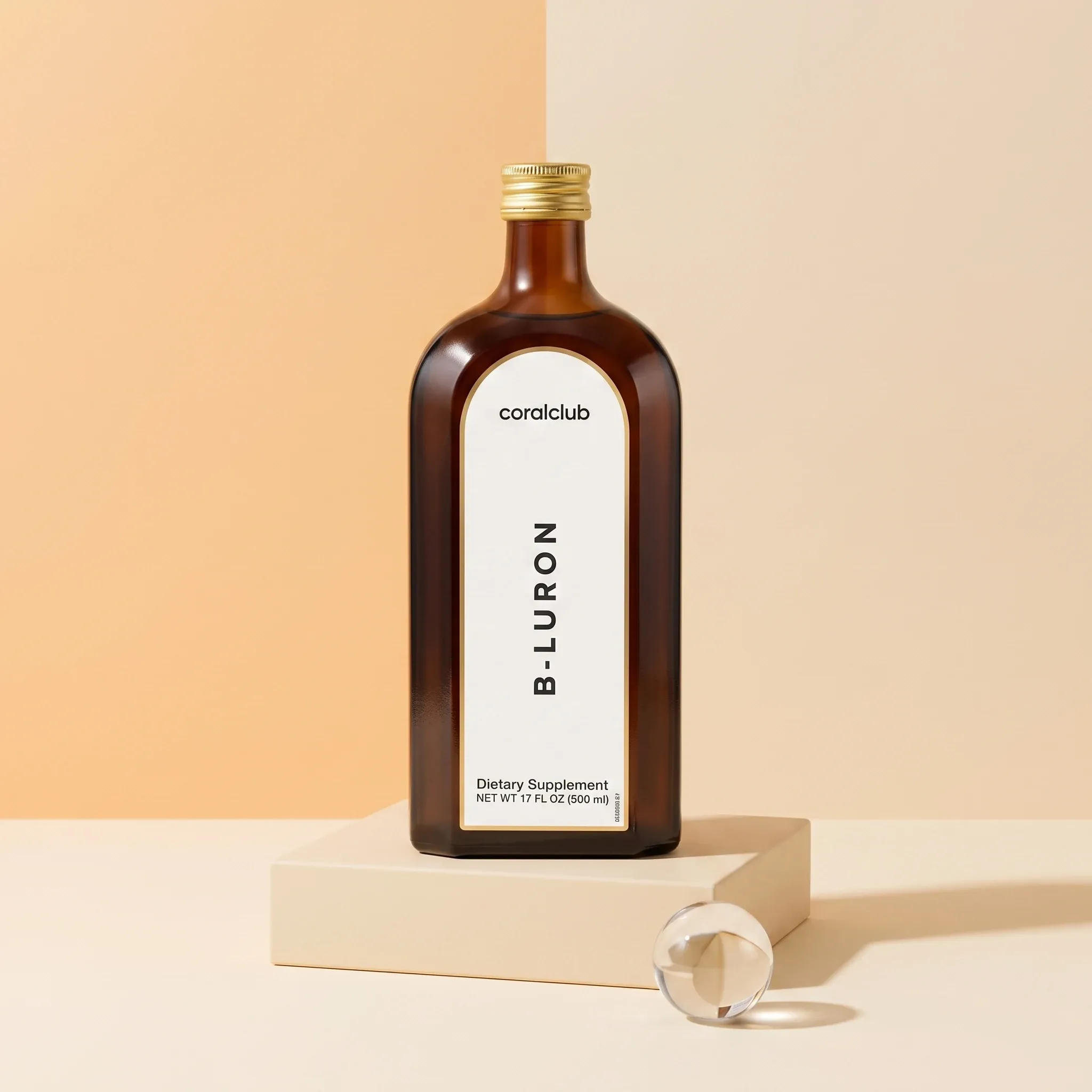 B-Luron 500 ml amber glass bottle on a cream plinth against a coral color-block background.